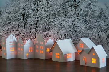 Close up of handmade small white cardboard houses with illuminated windows on snowy trees...