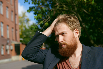 Fashion portrait of bearded hipster outdoor in sunny day. Handsome man in suit comb hair in the street. Blond Caucasian guy walk alone. Stylish look person.
