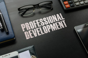 Black chalkboard with business accessories (notepad, magnifying glass, fountain pen, tablet, phone, glasses and calculator) and text PROFESSIONAL DEVELOPMENT. Top view.