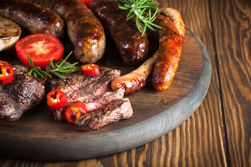 Close-up photo of mixed grilled meat platter. Beef, pork, poultry, sausages, grilled garlic, chili pepper, red tomatoes on wooden rustic background.