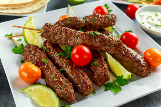 Homemade Kofta kebabs on skewers with pita, lime, vegetables, sweet chili and yogurt sauce on white plate.