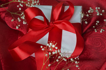 Gift in white packaging red ribbon tied on a red cloth. White fl