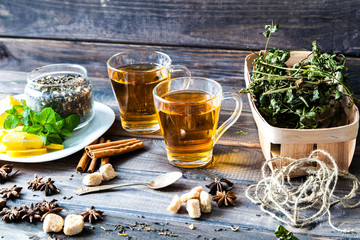 Two clear glasses of hot black herbal tea with lemon, mint leaves on old dark grey rustic wooden table. Summer, Autumn, winter drink. Dried herb in birch basket. Cinnamon for decoration. Low key.
