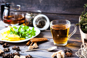 Glass and teapot of hot black herbal tea with lemon, mint leaves on dark grey rustic wooden table. Summer, Autumn, winter drink. Dried herb on old newspaper. Cinnamon for decoration.