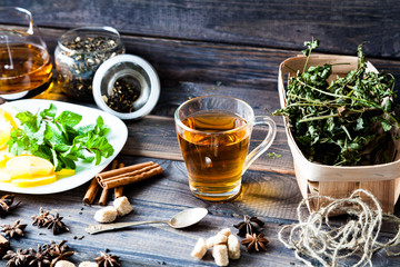 Glass and teapot of hot black herbal tea with lemon, mint leaves on dark grey rustic wooden table. Summer, Autumn, winter drink. Dried herb on old newspaper. Cinnamon for decoration.