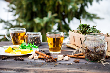 Two glasses of hot black herbal tea with lemon, mint leaves on old dark grey rustic wooden table. Summer, Autumn, winter drink. Dried herb in birch basket. Cinnamon for decoration. Fir tree background