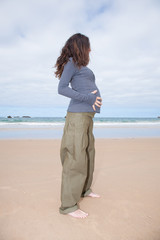 brunette brown hair pregnant woman with grey jersey green trousers and black sunglasses standing with hands on tummy in sand beach ocean in Asturias Spain
