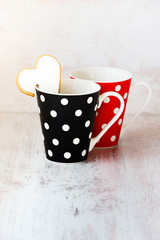 A pair of black and red  polka dotted coffee cups with a heart shaped homemade cookie on the edge over white wood background.