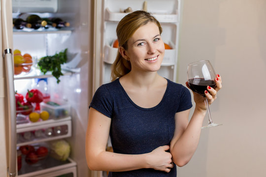 Young woman in the sleepwear drinking red wine near the refrigerator