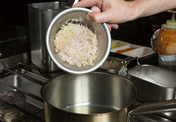 Minced garlic and finely sliced onion being dropped into a metal frying pan of olive oil. Step by step recipe cooking.