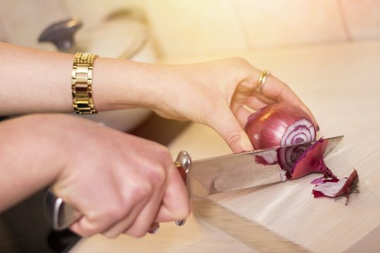 Close-up of a woman's hand cutting an onion on a wooden board. Lens flare in the background.