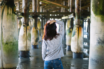 Brunette girl back in a gray sweater. Young women stands under a pier and looks at the ocean. Pacific beach pier