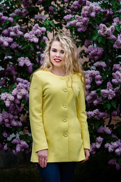 Blonde girl in a yellow coat stands against the backdrop of blooming lilacs in spring