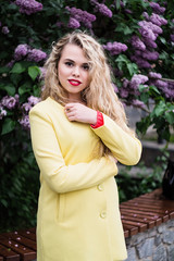 Blonde girl in a yellow coat stands against the backdrop of blooming lilacs in spring