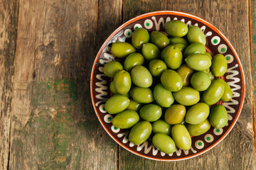 Green olives in brown bowl on wooden background. Top view. Copy space.
