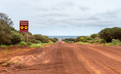Outback Red Gravel Road. Red gravel road near Kings Canyon, NT Australia. Reduce speed sign with iconig kangaroo warning. Cloudy sky, red ocher soil.