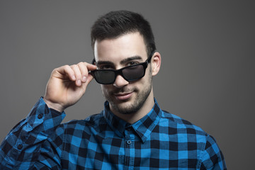 Obraz premium Seductive young man taking off sunglasses smiling and looking at camera over gray studio background. 