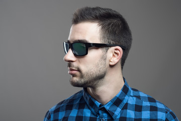 Fototapeta premium Profile view of young bearded man in plaid shirt wearing sunglasses looking away over gray studio background. 