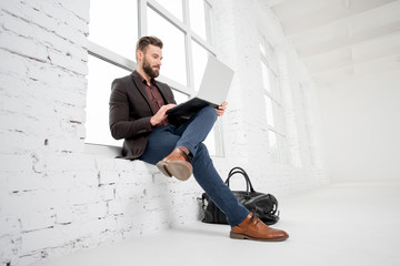 Elegant businessman sitting on the window with laptop in the white office interior