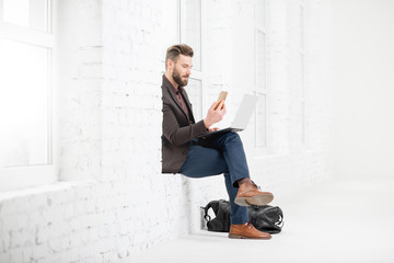 Elegant businessman sitting on the window with laptop and mobile phone in the white office interior