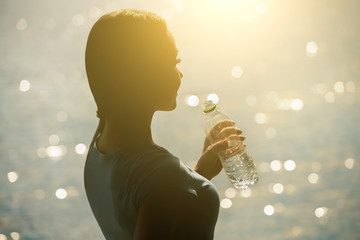 Silhouette of a young female athlete in tracksuit drinking water from a bottle on the beach in summer during morning exercises. Sport and healthy lifestyle.