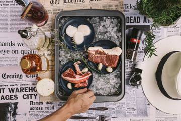 Adjusting Beef, Pork, and Scallop Preparing for Cooking on Iced Plate with Olive Oil, Wine, Half of Onion and Black Pepper on Newspaper with Plant and Hat.