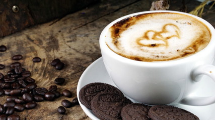 Cappuccino Coffee and sweet chocolate cookies. A cup of latte, cappuccino or espresso coffee with milk put on a wood table with dark roasting coffee beans. Drawing the foam milk on top.