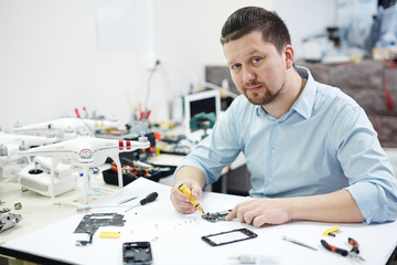 Portrait of modern man in workshop looking at camera while repairing electronics, smartphones, drones, laptop on table with different tools
