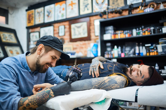 Happy young tattooer in ball-cap working with client