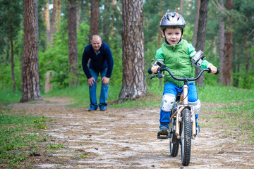 Naklejka premium Little kid boy of 3 years and his father in autumn forest with a bicycle. Dad teaching son. Man happy about success. Child helmet. Safety, sports, leisure kids concept