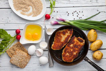 Fried chicken fillets in breading, with ingredients on white wooden table