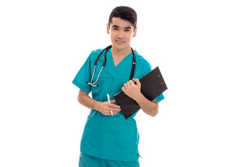cheerful young brunette male doctor with stethoscope in uniform looking at the camera and make notes isolated on white background