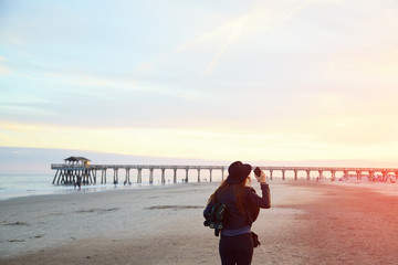 Woman in hat holding cell phone on beach. Girl walking on coast with smartphone in her hand. Female in black clothes using mobile phone near ocean pier jetty.