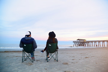 Couple sitting in deck chairs at beautiful sunset beach. Man and woman in hats and casual clothes relaxing near ocean pier jetty. Peaceful scene, calming waves, pastel cloudy sky, coast, wet sand.