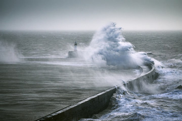 A monster wave crashes over a sea wall near a lighthouse during a storm