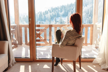Woman sitting in a comfortable chair drinking coffee and looking through window at snow covered mountain.