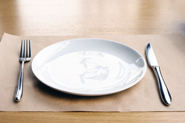 Empty white plate, fork and knife, on a wooden table in a restaurant