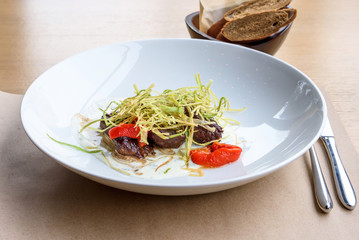 Beef medallions with greens, fry, red boiled pepper and white cheese sauce on a white plate, brown bread, restaurant table