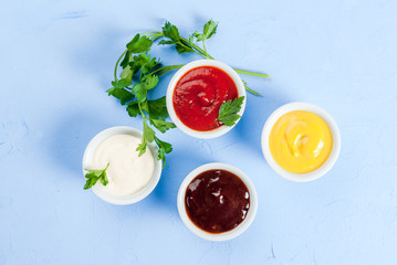 Classic set of sauces in white saucers: American yellow mustard, ketchup, barbecue sauce, mayonnaise. On cutting board light blue stone concrete table top view, copy space