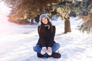 Beautiful young girl sits on to snow in warm clothes in winter outdoors