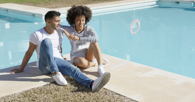 Young woman with an afro hairstyle relaxing with her boyfriend poolside on a hot summer day sitting together chatting on the tiled surround