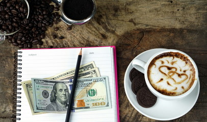 Cappuccino coffee and sweet chocolate cookies, US bills money. A cup of latte, cappuccino or espresso coffee with milk put on a wood table with dark roasting coffee beans.