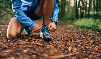 Fit male jogger ties shoes while day training for cross country forest trail race in a nature park.