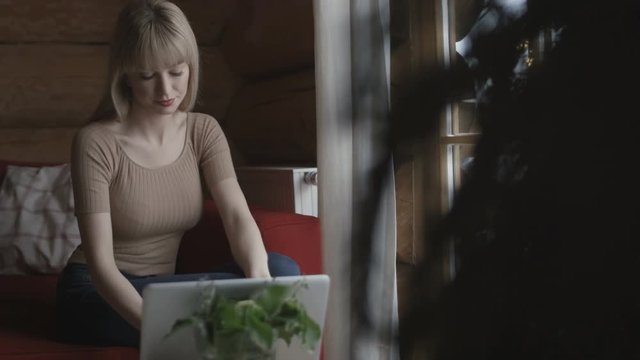 Young pensive woman sitting on the couch while typing and reading emails on a laptop computer with winter background on the window. Shot on Red Epic.