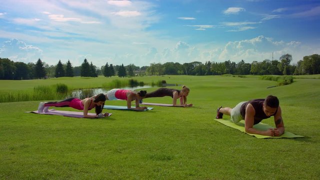 Group of people doing plank exercise on green grass at sunny day. Outdoor fitness class in park. Women group fitness outdoor with fitness trainer. Group fitness people exercising in spring park