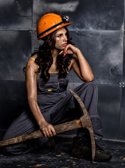 sexy female miner worker with pickaxe, in coveralls over his naked body, sitting on the floor on backdrop of steel wall