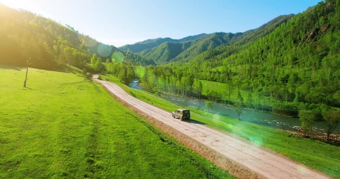 UHD 4K aerial of vehicle driving towards the sunrice. Mid-air shot around a dirty suv retro car driving on green countryside rural road.