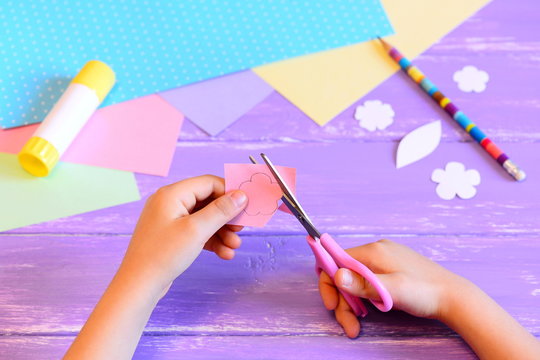 Small child creates a greeting card for mom. Step. Child holds scissors and cuts a flower from paper. Materials for creative art on a wooden table. Crafts idea for Mother's day or birthday or March 8