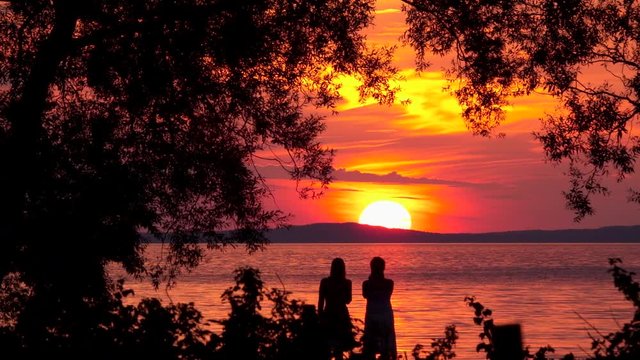 Silhouette of two people watching orange sunset over lake. Beautiful nature lifestyle travel background. Summer night in Sweden, Scandinavia. 