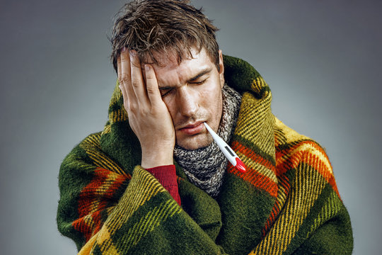 Young man suffering from a headache. Photo of sick man wrapped in blanket with a thermometer in his mouth.
